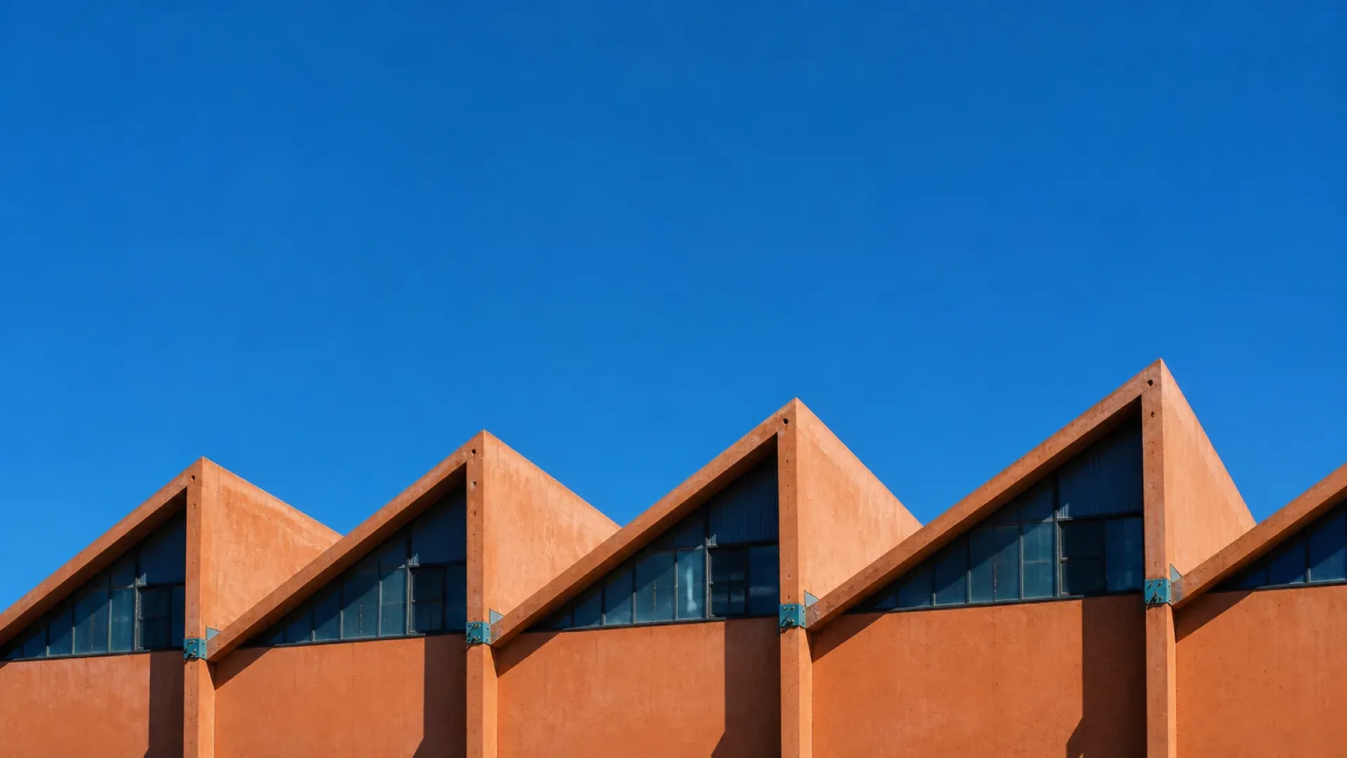 A repeating sawtooth factory roofline in terracotta against a clear blue sky
