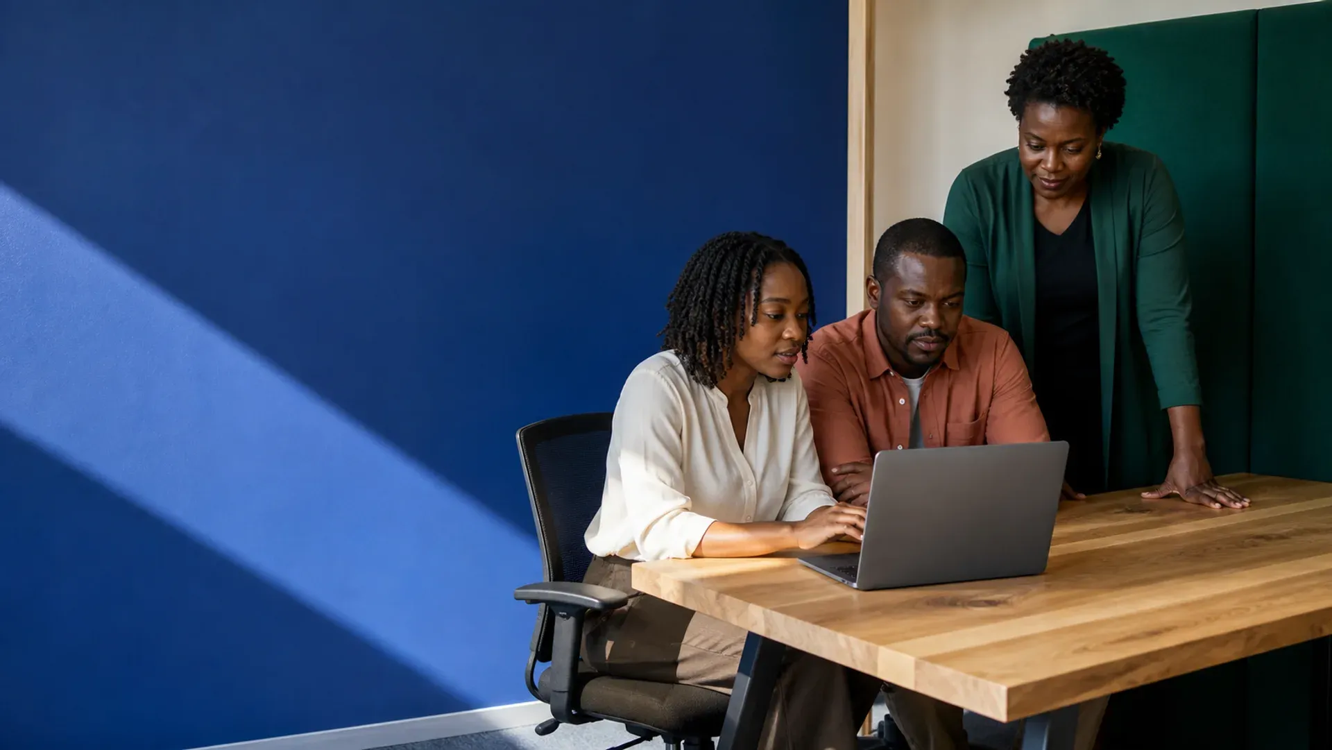 Three colleagues gathered around a laptop at a wooden table against a cobalt wall
