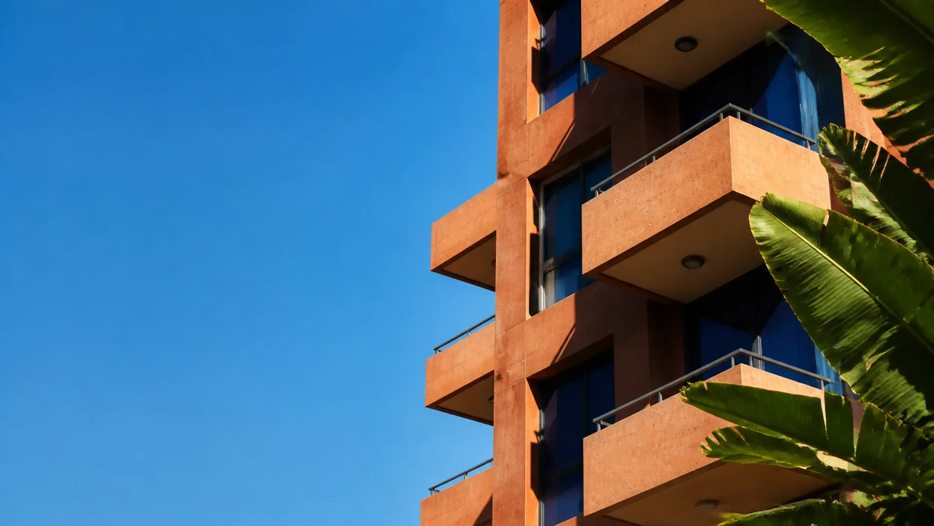 A modernist tower with stepped balconies and palm fronds against a blue sky